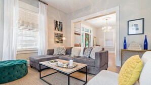 A modern living room with a grey sectional sofa, a green ottoman, and a white armchair. There is a coffee table with magazines and a candle. The room has a large window with white curtains, a chandelier, and artwork on the walls. - The Glen Iris Getaway (Birmingham)