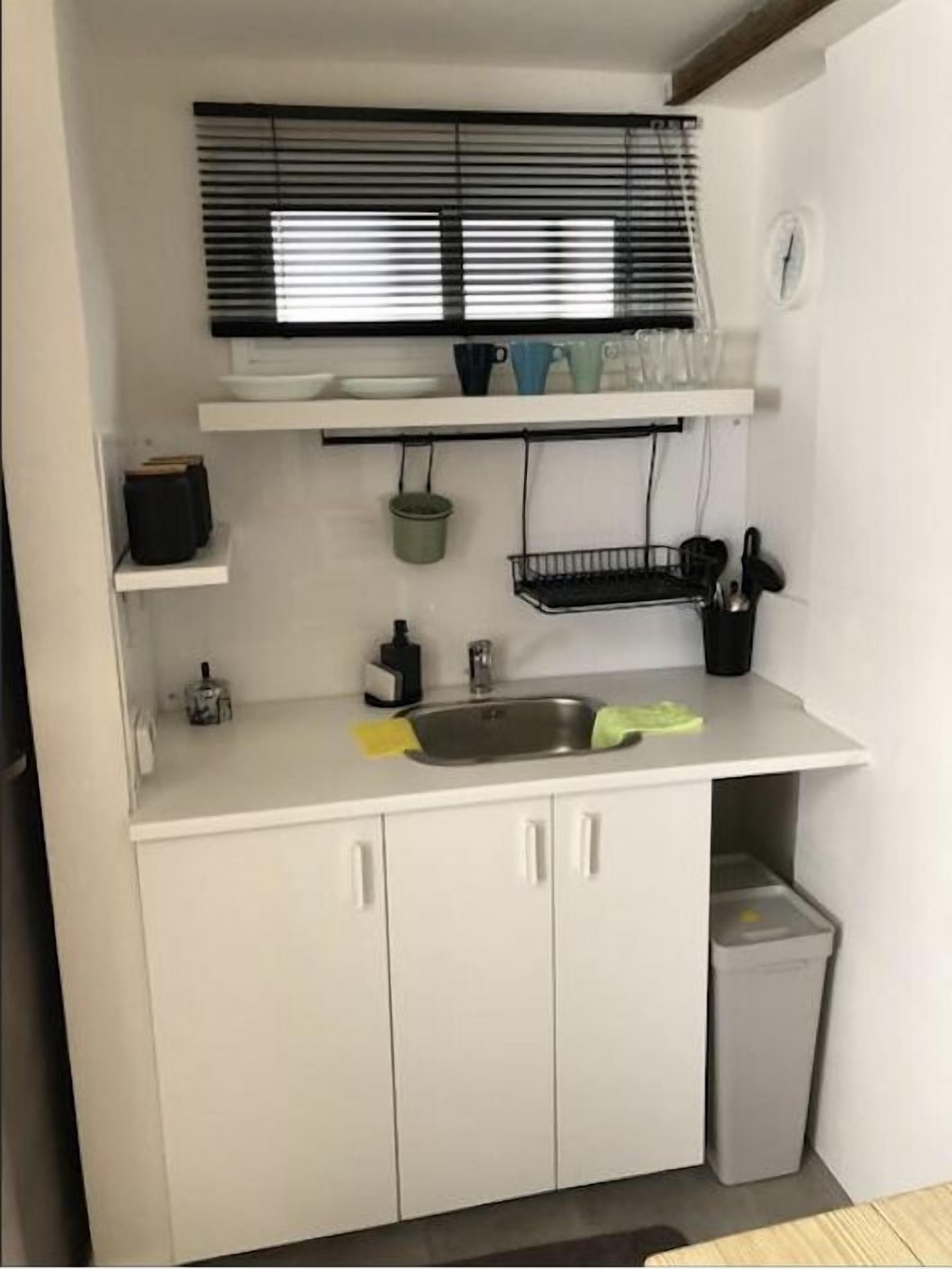 A compact kitchen with a sink, a window with blinds, a small trash bin, and a few shelves with kitchenware.