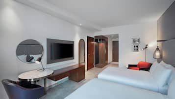 A modern hotel room with a large bed, a desk with a round mirror, a flat-screen TV, and a red chair.