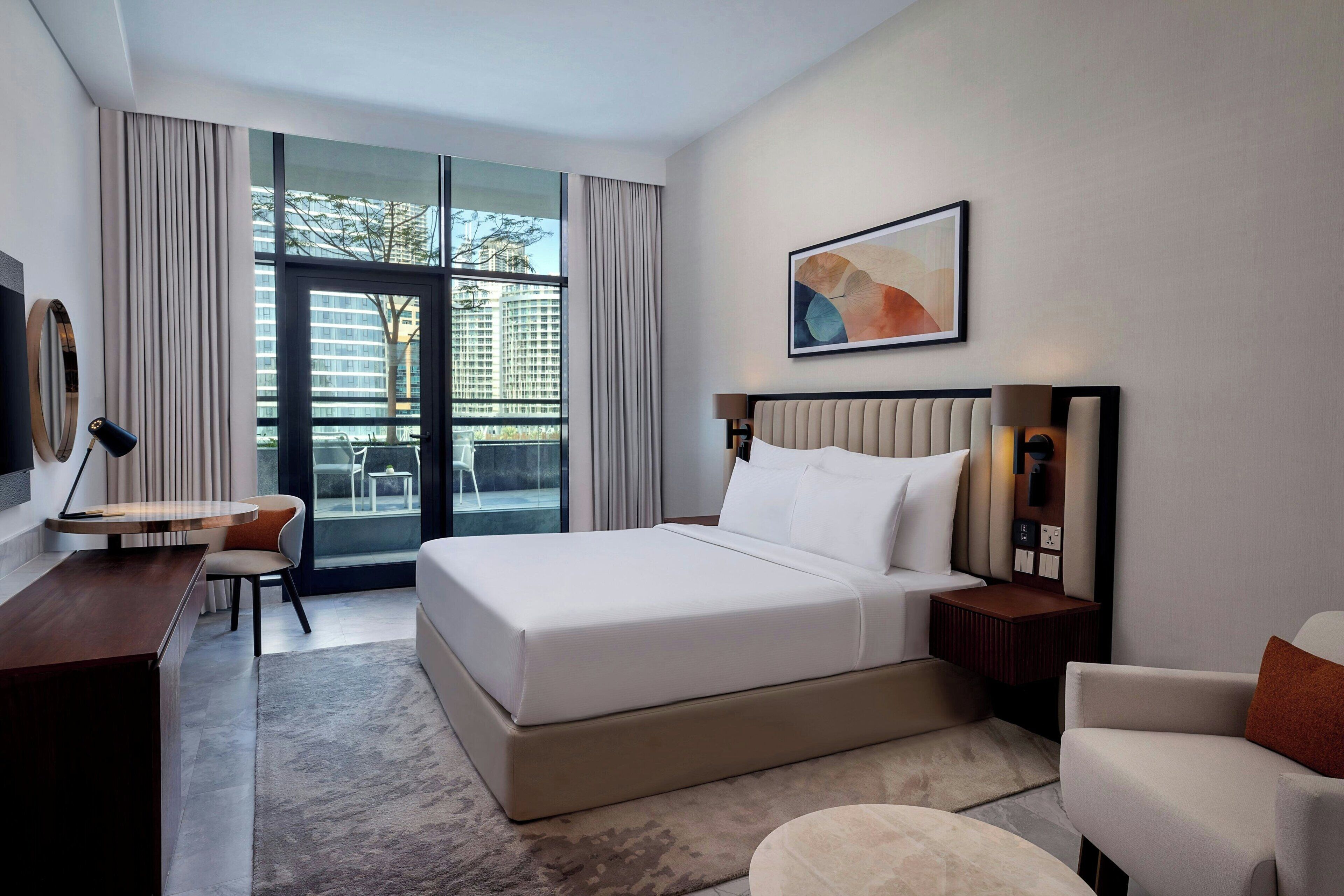 Une chambre d’hôtel moderne dotée d’un grand lit, d’un bureau, d’une chaise et offrant une vue sur la ville grâce à de grandes fenêtres.