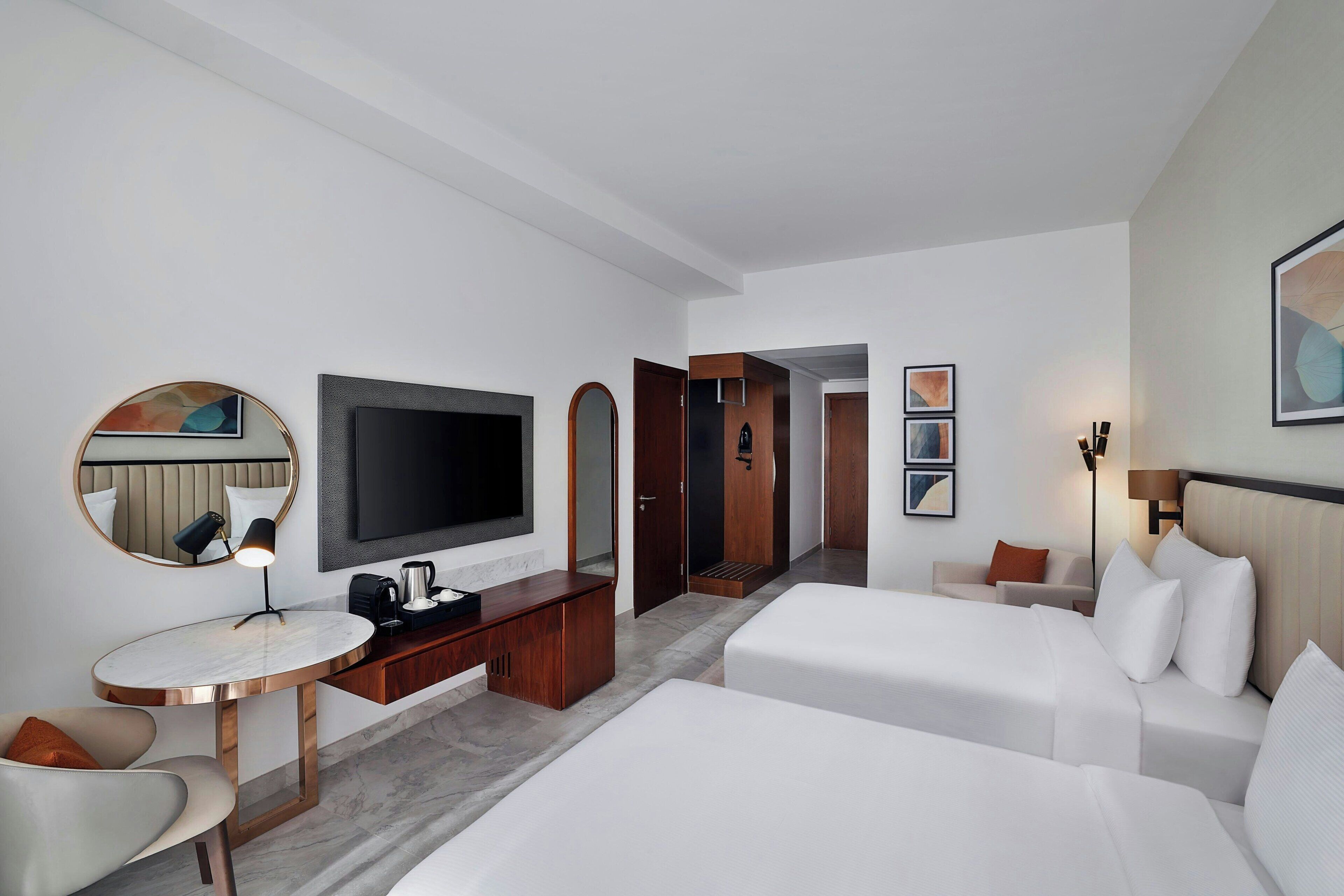 Een hotelkamer met een televisie, bureau, stoel, twee bedden, een garderobe en een deur naar een andere kamer.