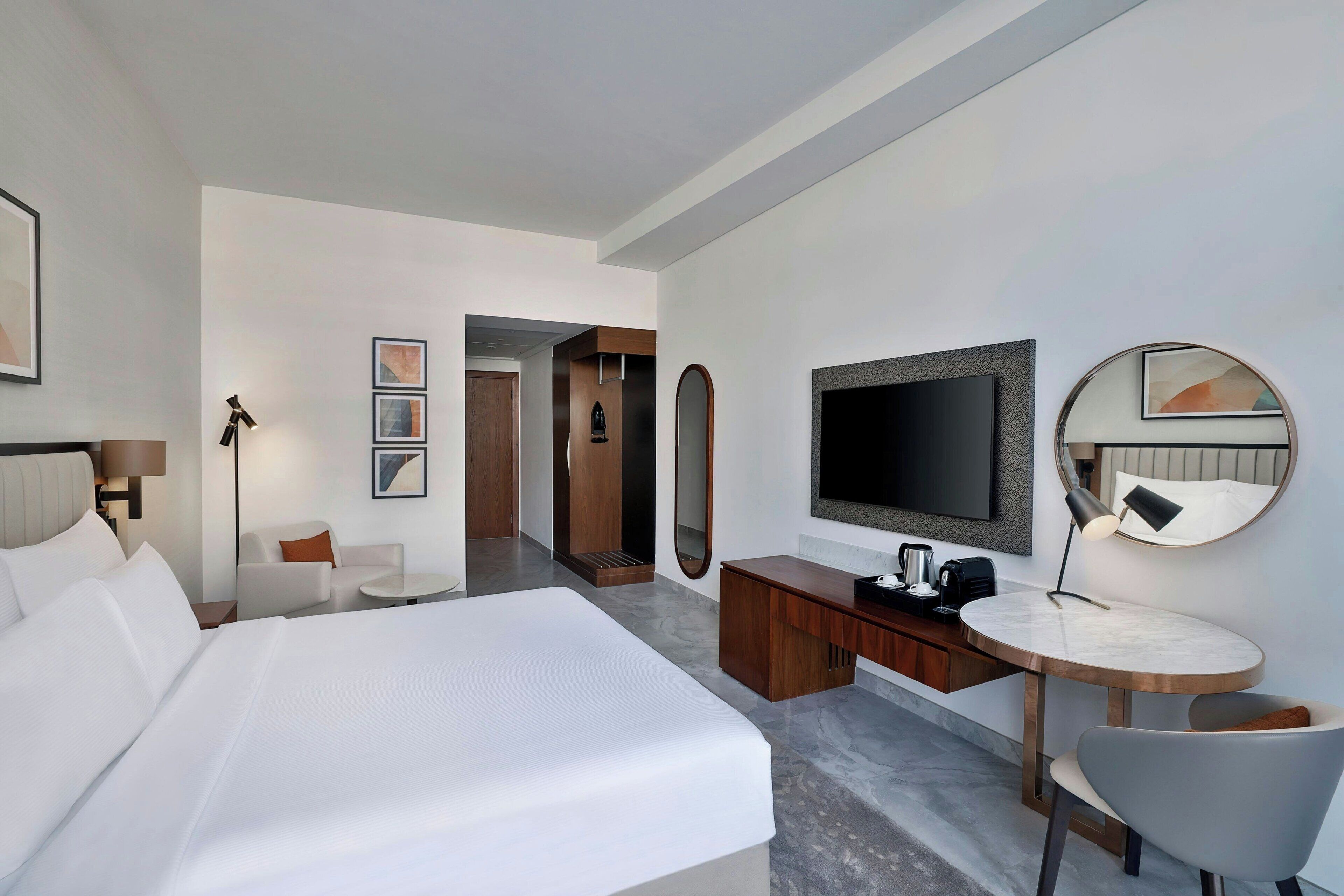 A modern hotel room with a large bed, a desk with a flat-screen TV, a round mirror, and a seating area with a chair and a small table.