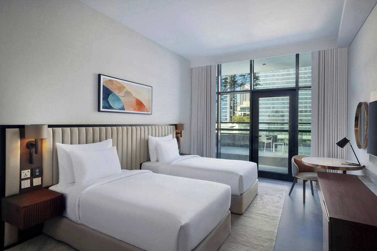 A hotel room with two beds, a desk, a chair, a large window with a view of buildings, and a framed abstract painting on the wall.