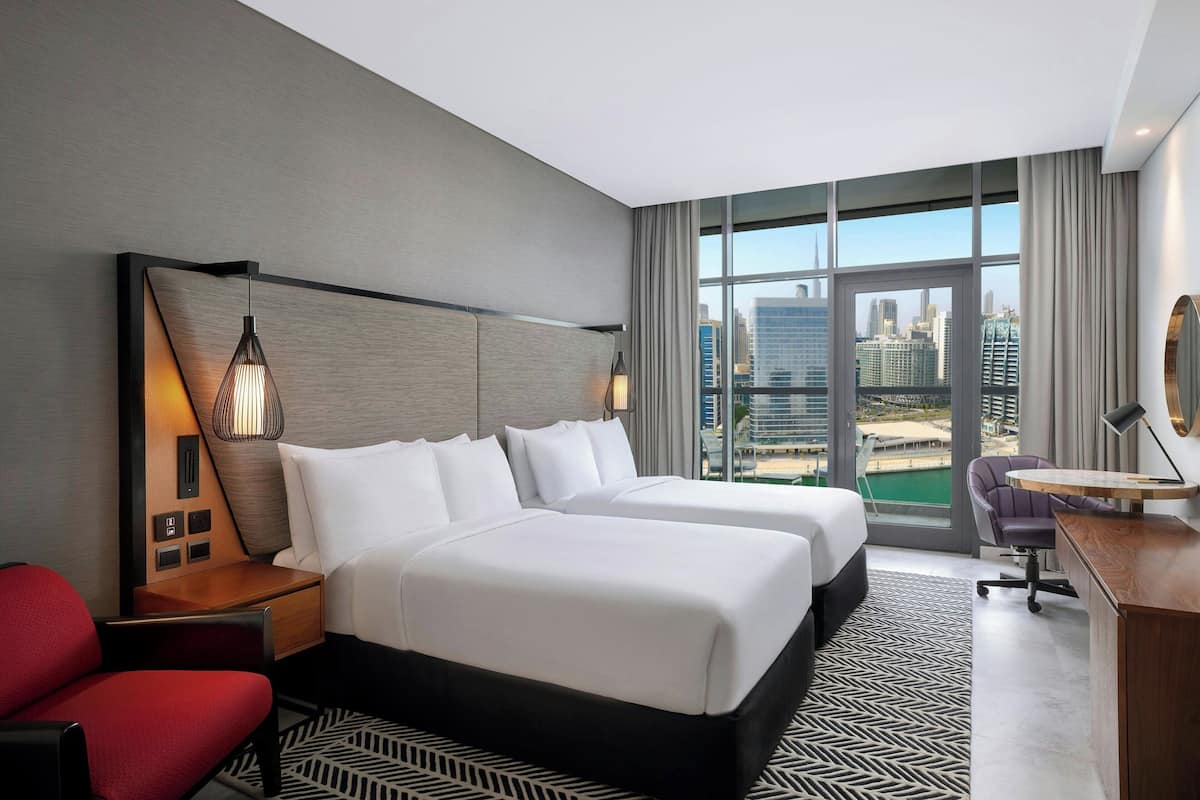 A hotel room with two beds, a desk, a chair, and a large window offering a city view.