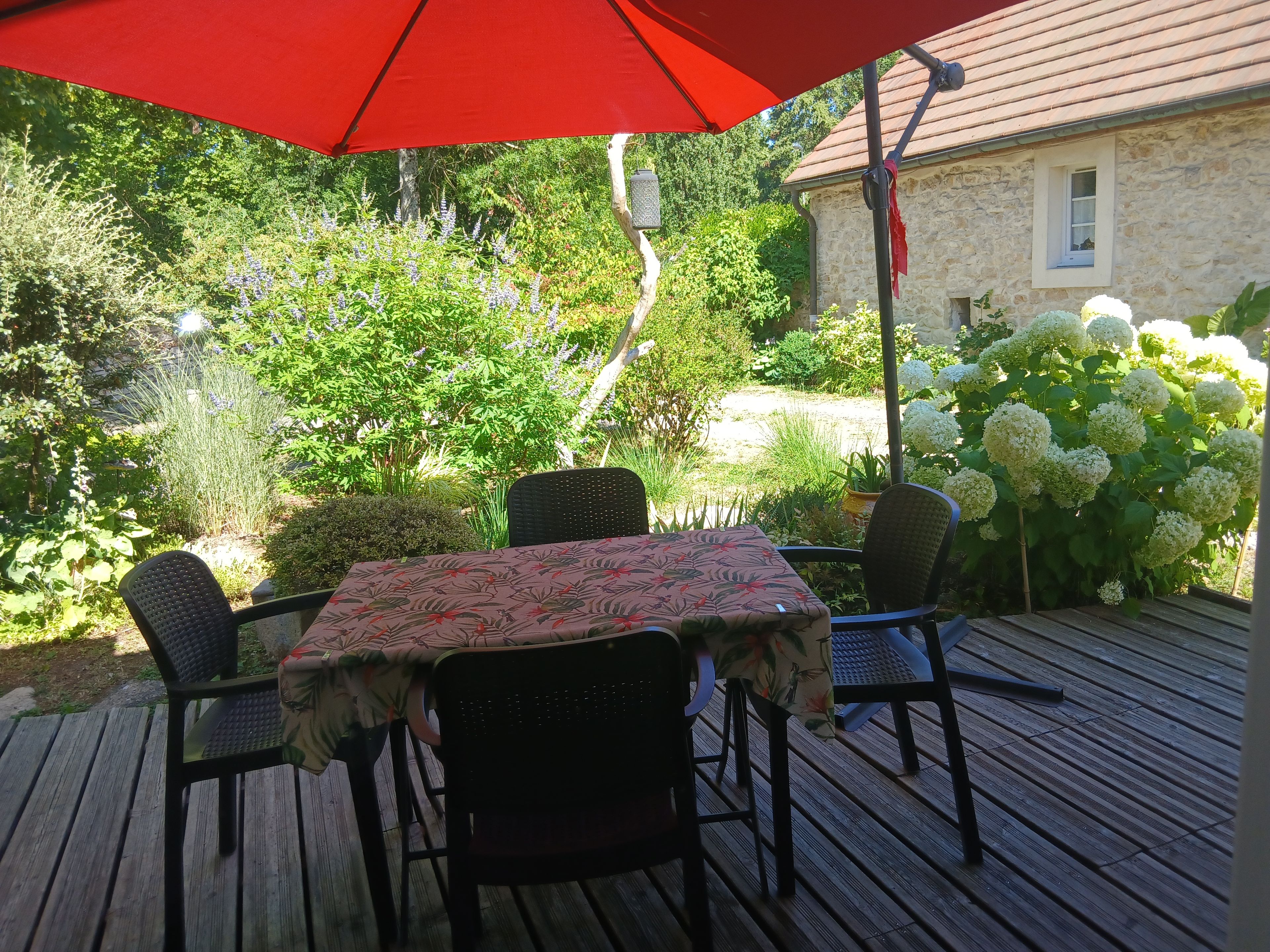 En udendørs terrasse med et bord og stole, omgivet af frodigt grønt og en stengård.