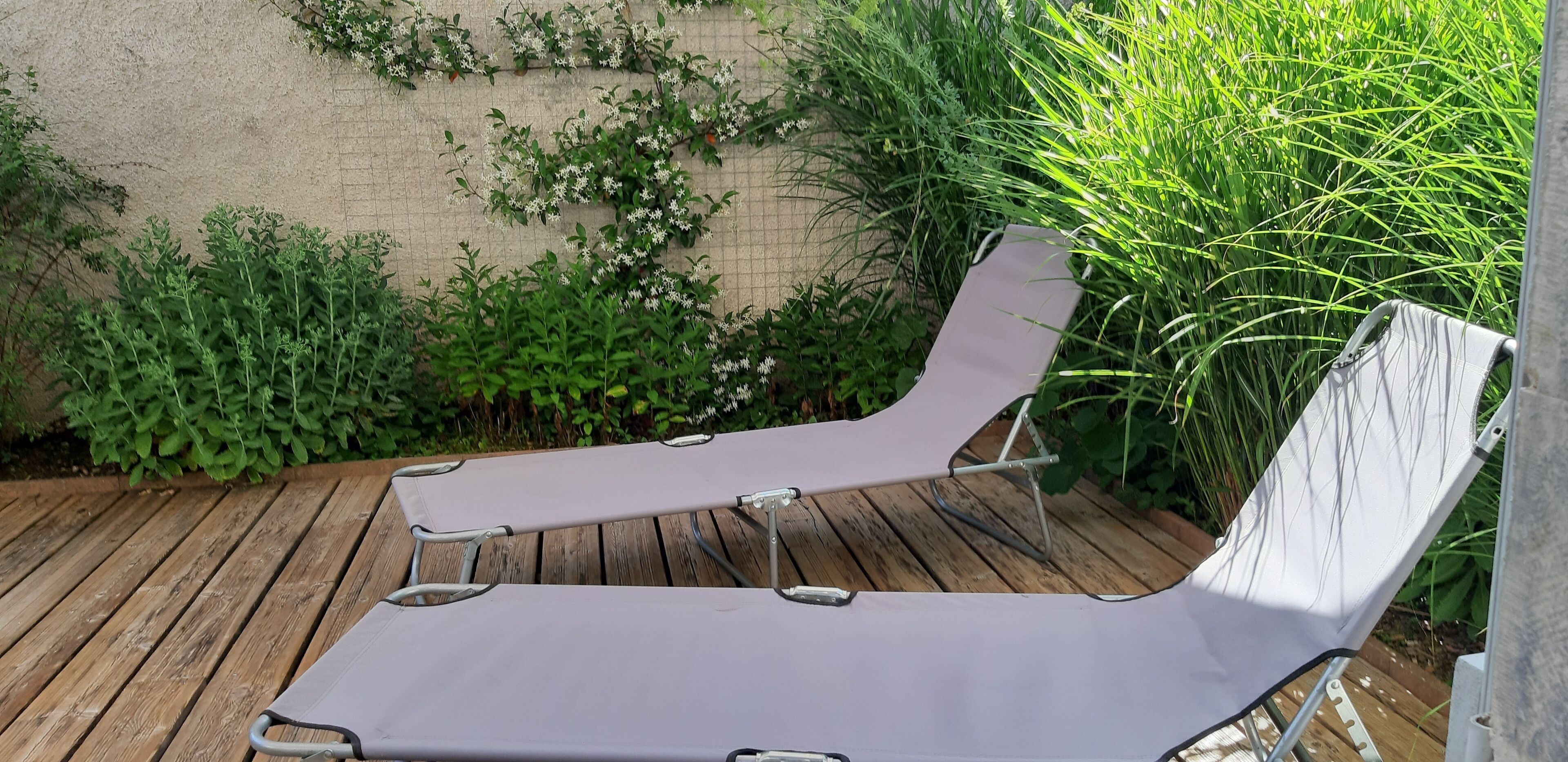 Twee ligbedden op een houten terras, met een bakstenen muur en weelderige begroeiing op de achtergrond.