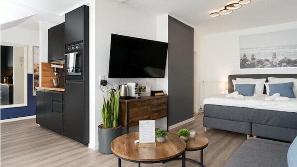 A modern hotel room with a kitchenette, a flat-screen TV, a bed with blue pillows, and a small dining area.