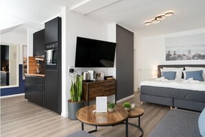 Een moderne hotelkamer met een kitchenette, een flatscreen televisie, een bed met blauwe kussens en een klein zithoekje.