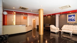 Un área de recepción con un mostrador de recepción identificado como "B2" y un cartel que dice "RECEPTION", un gran pilar dorado y dos zonas de estar con sillones de cuero.