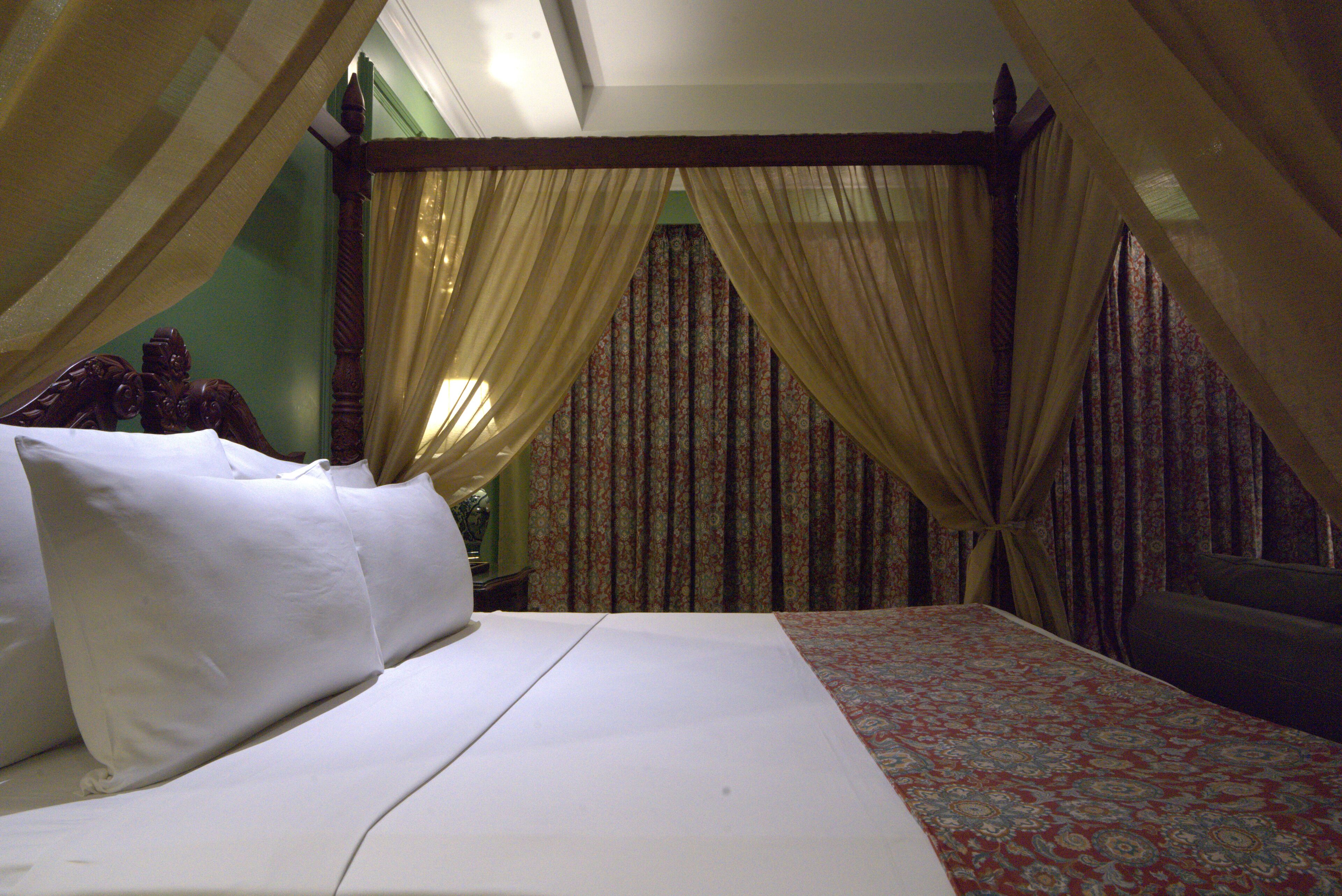 A bed with white linens and a patterned bedspread, a wooden headboard, and sheer curtains.