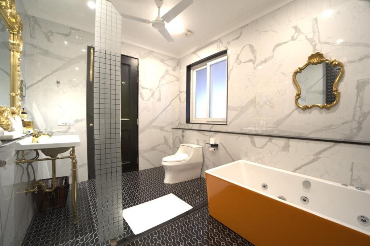 A bathroom with a marble wall, a white toilet, a bathtub, a gold-framed mirror, and a ceiling fan.