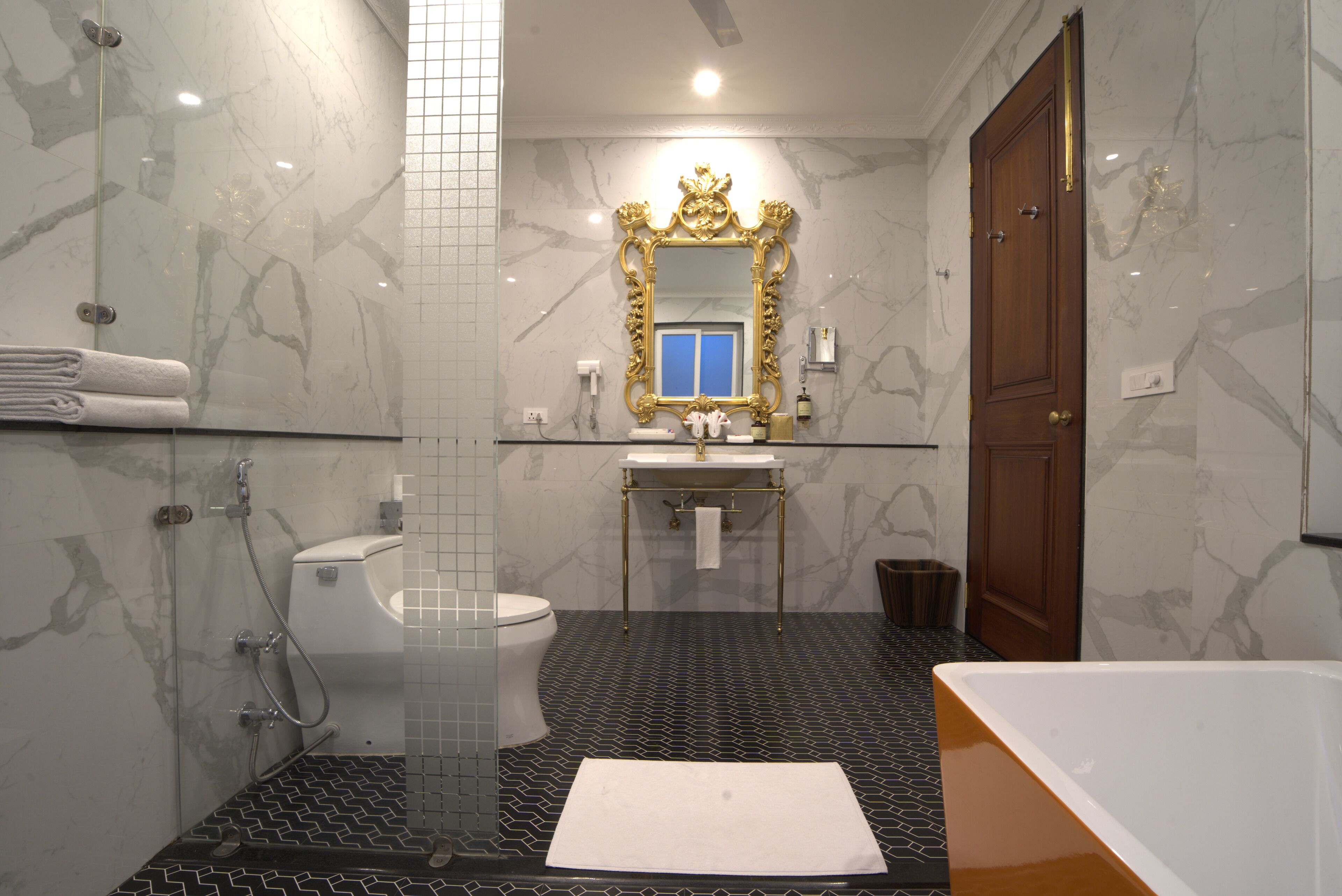 A modern bathroom with a glass-enclosed shower, a white toilet, a gold-framed mirror, and a wooden door.