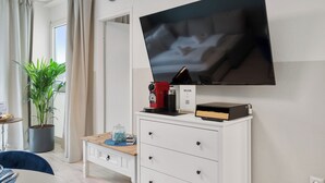 A modern living room with a flat-screen TV mounted on the wall, a coffee machine, a potted plant, and a small table with a blue mat. - Superior 2-zimmer 36qm Kingsize Boxspring 55' TV Home-office 11min Hbf (Duisburg)