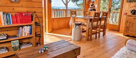 Interior de uma cabana de madeira com área de jantar, estantes de livros e vista para o exterior.