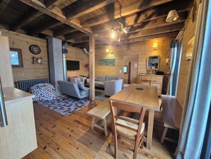 Ein gemütliches Blockhäuschen-Interieur mit Holzboden, einem Esstisch mit Stühlen, einer Couch, einem Heizkörper und einer Uhr an der Wand.