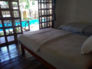 A room with a bed, a window with a view of a pool and greenery, and a wooden floor. - Kokomo Dive Resort (Mambajao)