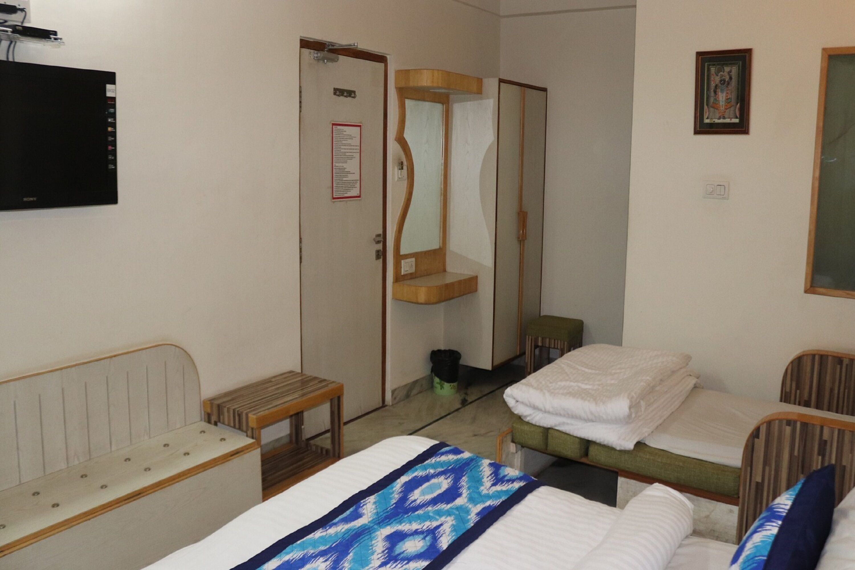A hotel room with two beds, a wooden bench, a small table, a mirror, and a television mounted on the wall.
