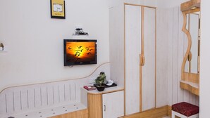 A room with a wooden bench, a wardrobe, a small stool, a television mounted on the wall, and a potted plant on the bench. - Vrinda Hotel (Nathdwara)