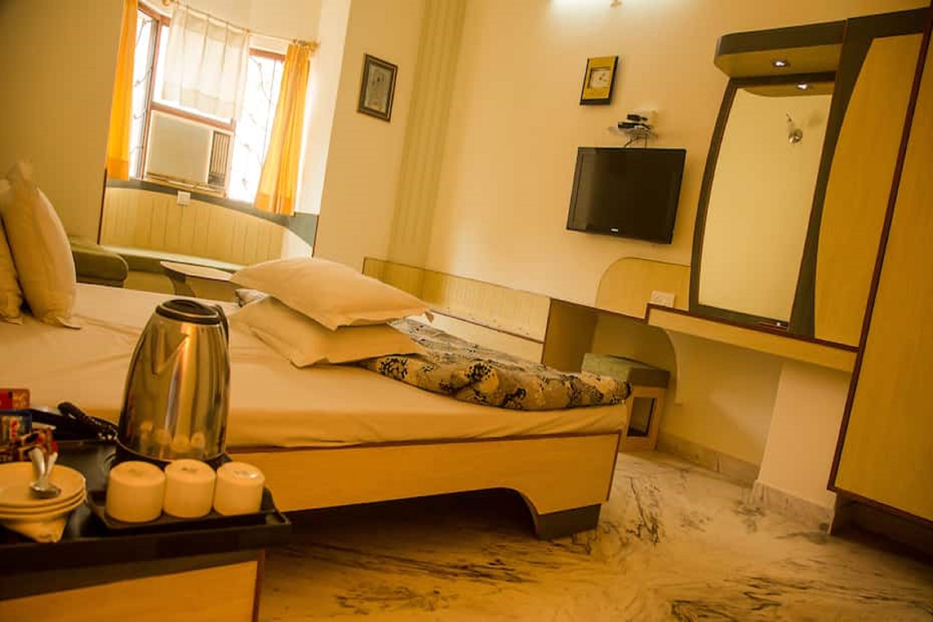 A hotel room with a bed, a bedside table with a kettle, a mirror, and a television.