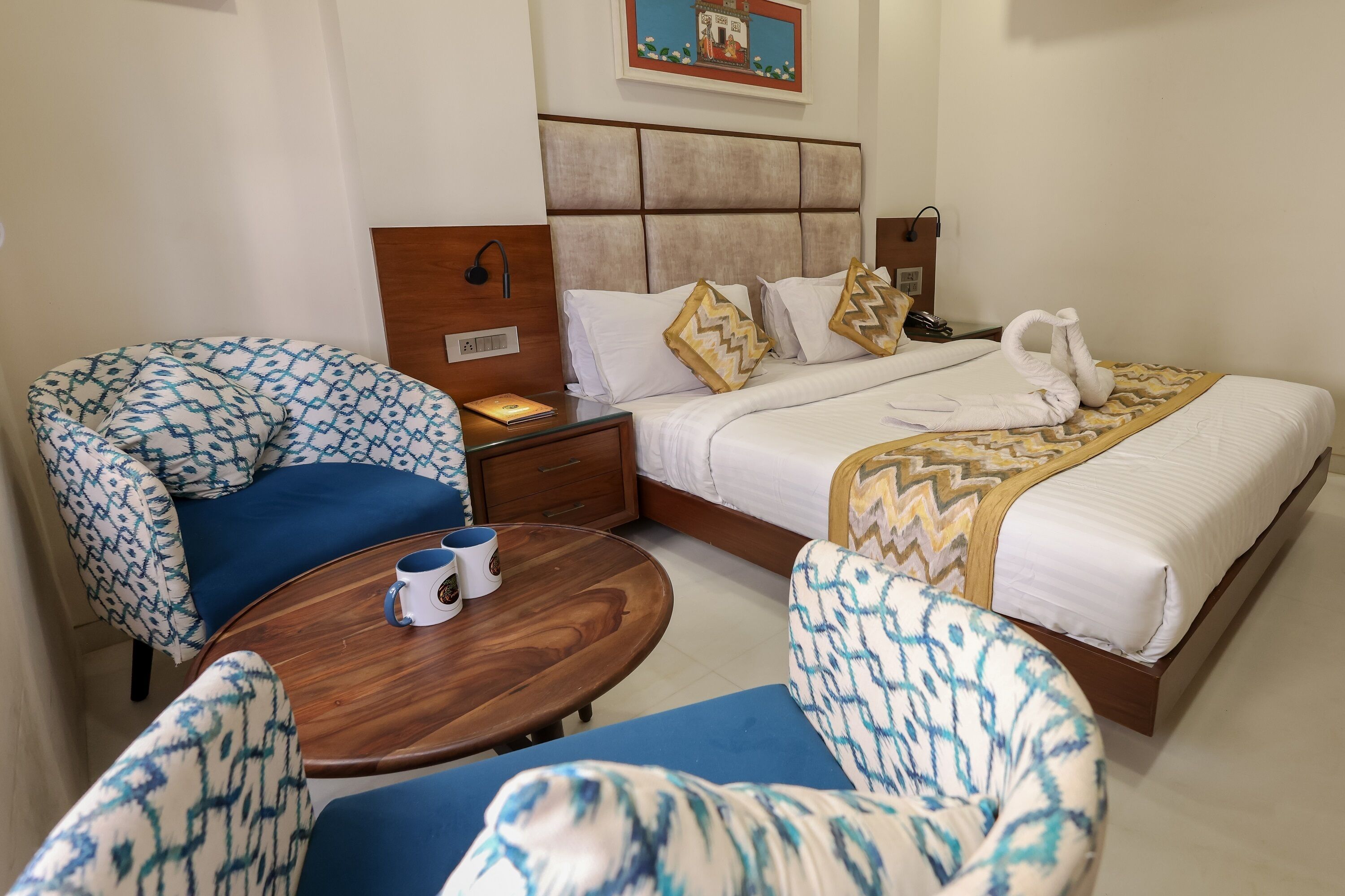 A hotel room with a bed, two patterned armchairs, a small wooden table with a mug, and a nightstand with a lamp.
