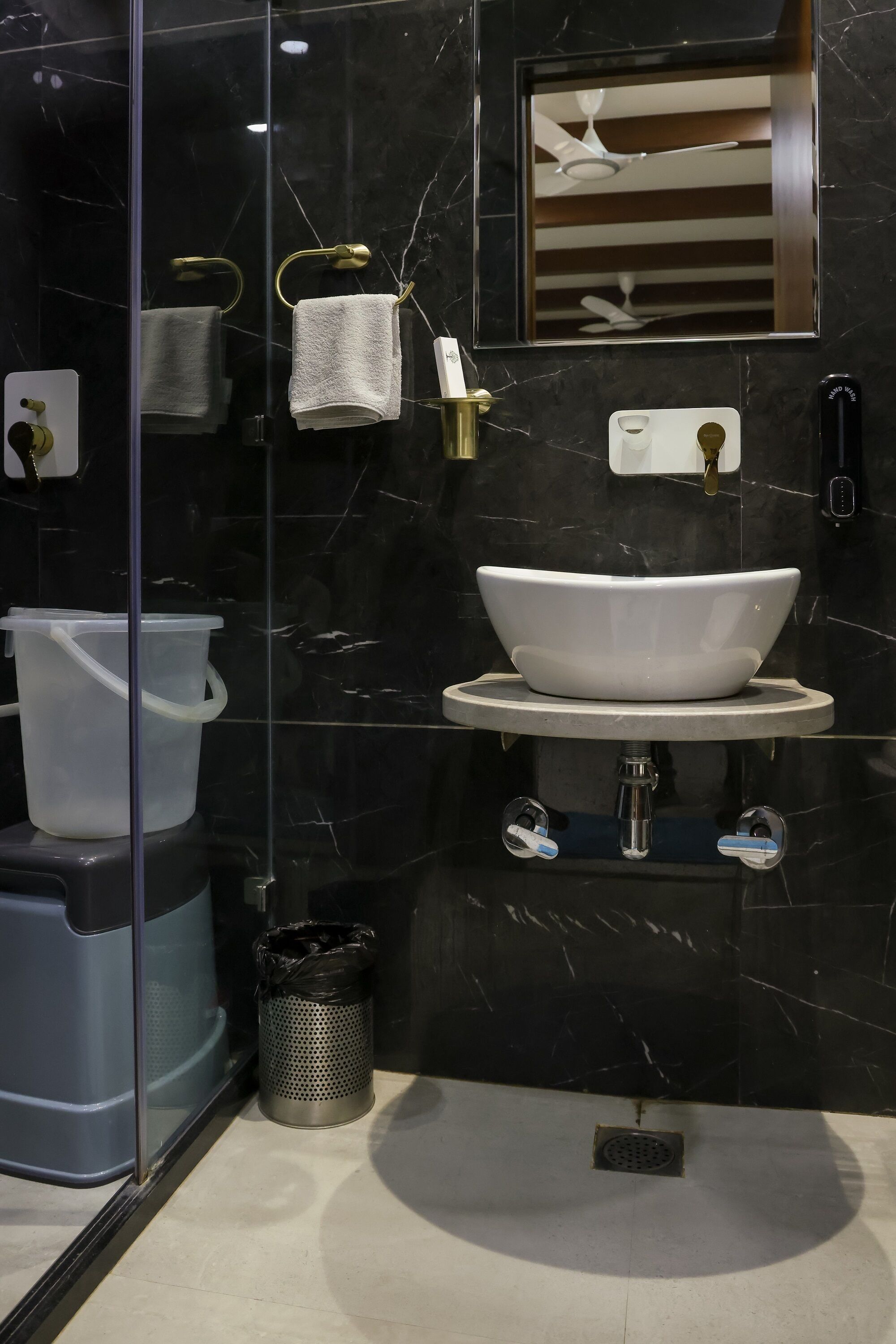 A bathroom with a white vessel sink, a glass shower enclosure, a black marble wall, a small trash bin, and a towel rack.