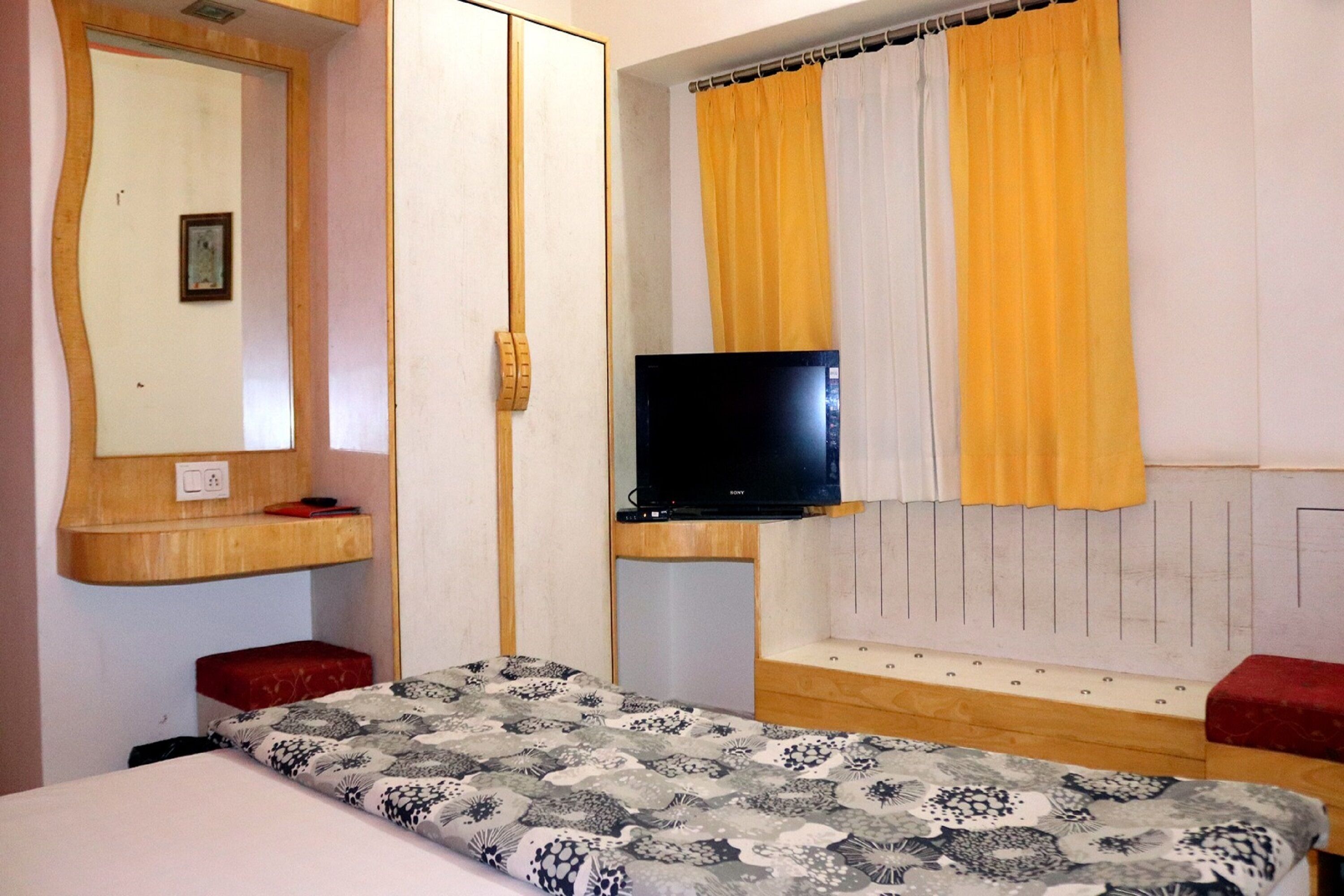 Un dormitorio con cama, televisión, espejo y ventana con cortinas amarillas.