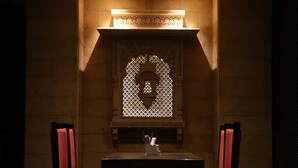 A dimly lit room with a table and two chairs, a decorative window, and a patterned wall. - Vrinda Hotel (Nathdwara)
