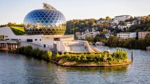 A modern building with a large spherical glass dome, situated by a river with a small island in the foreground.