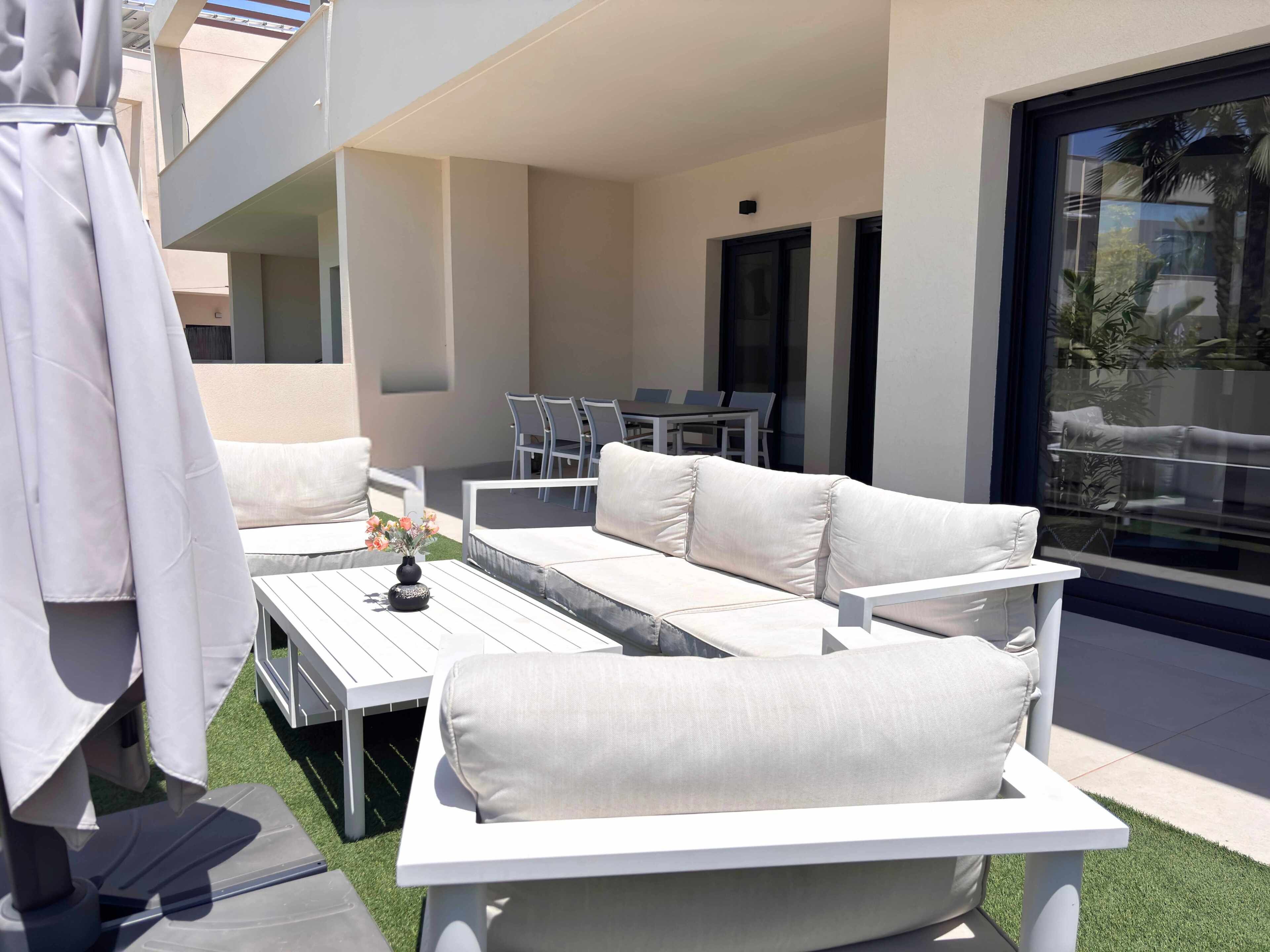 Un área de estar exterior moderna con un sofá blanco, una mesa, sillas, un pequeño jarrón decorativo y un vidrio que da a un balcón.