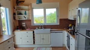 Una cocina con armarios blancos, un refrigerador de acero inoxidable, una ventana con vistas a follaje y una lámpara colgante.