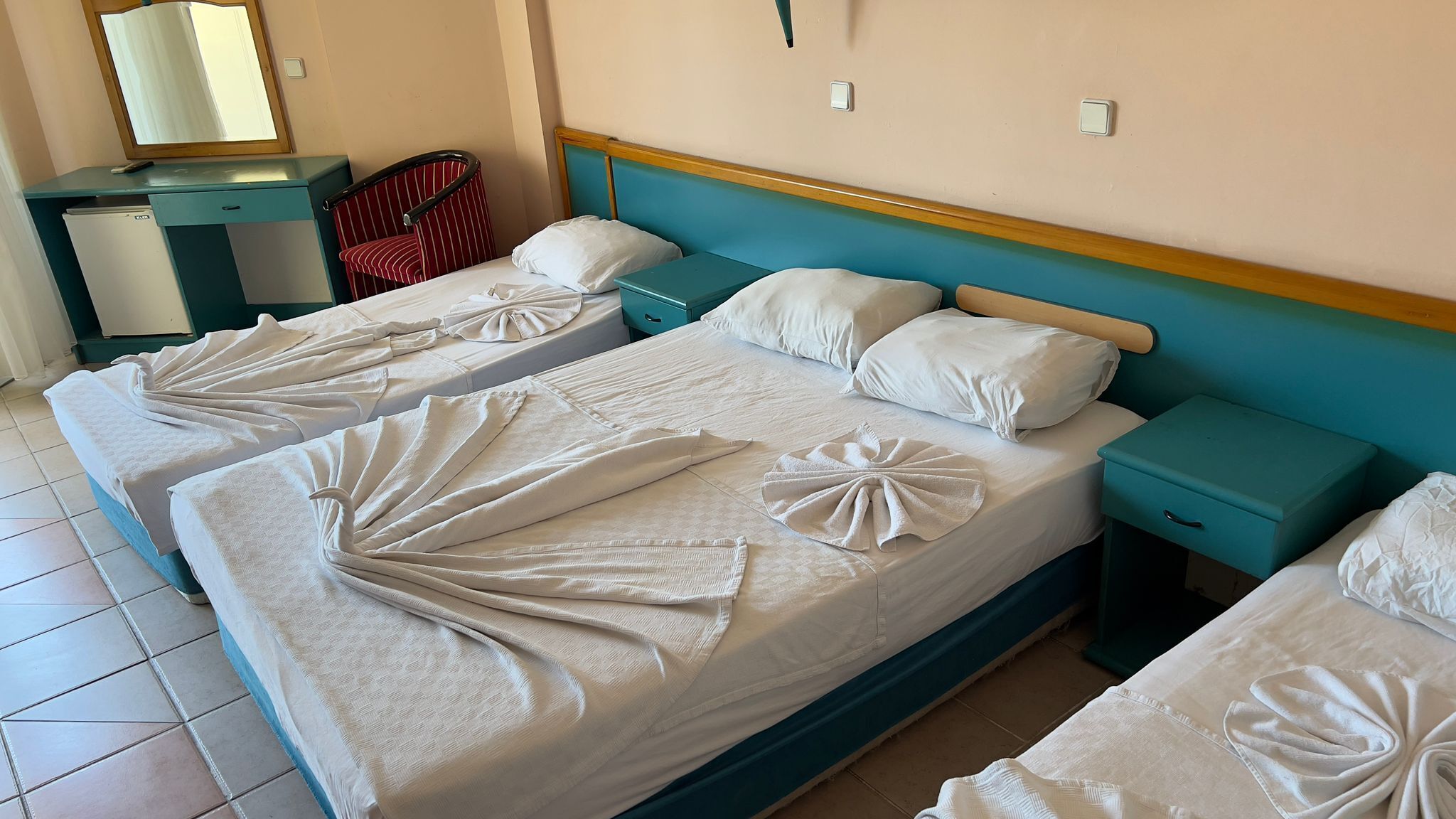 A hotel room with two beds, each with towels arranged in a fan shape. There is a small desk with a mirror and a chair, and a nightstand with a lamp.