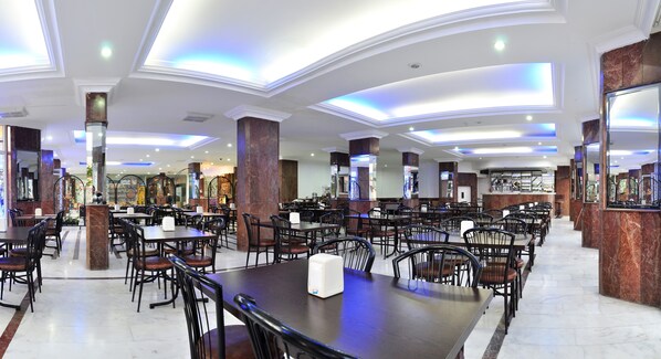 Un restaurante espacioso con techos altos, columnas de mármol y un diseño moderno. Las mesas y sillas están ordenadas y la iluminación es cálida y acogedora.