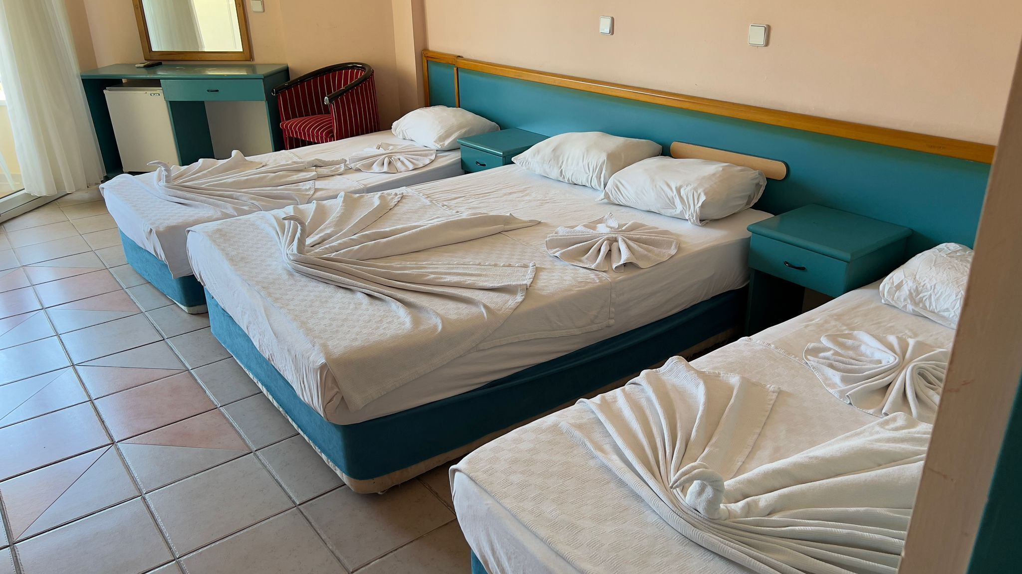 A hotel room with three beds, each with towels arranged in a fan shape. There is a small desk with a mirror and a red chair.