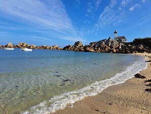 A coastal scene with clear water, a sandy beach, and a lighthouse on a rocky outcrop. - Maison Entièrement Rénovée ,à 600m de la mer Ideale Pour Passer de Bons Moments! (Plounéour-Brignogan-plages)