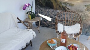 A cozy room with a white sofa, a wicker chair, a small table with snacks, a bottle of wine, and a view of the sunset over the beach. - Maison Entièrement Rénovée ,à 600m de la mer Ideale Pour Passer de Bons Moments! (Plounéour-Brignogan-plages)