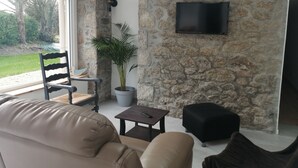 A modern living room with a stone wall, a flat-screen TV mounted on it, a leather sofa, a wooden chair, a small table, and a potted plant. - Maison Entièrement Rénovée ,à 600m de la mer Ideale Pour Passer de Bons Moments! (Plounéour-Brignogan-plages)