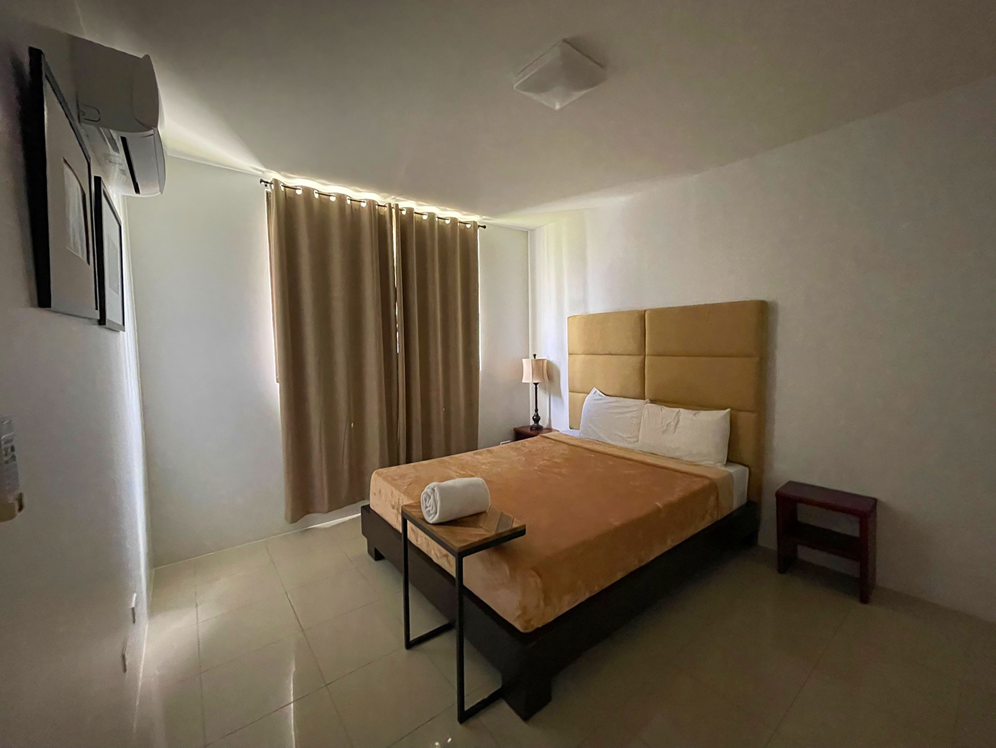A hotel room with a bed, a bedside table, a lamp, a TV mounted on the wall, and a window with curtains.
