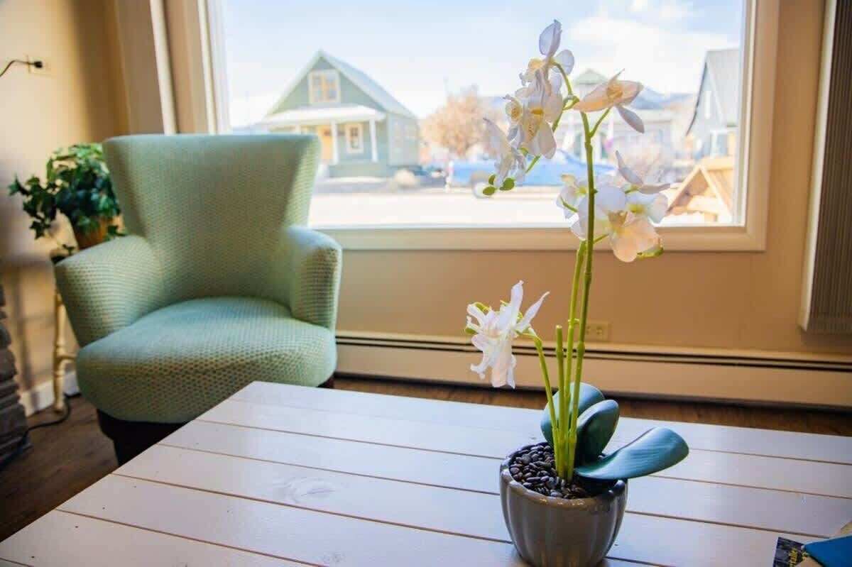 Ett rum med ett träbord, en orkidé i en kruka, en grön fåtölj och ett fönster med utsikt över hus.