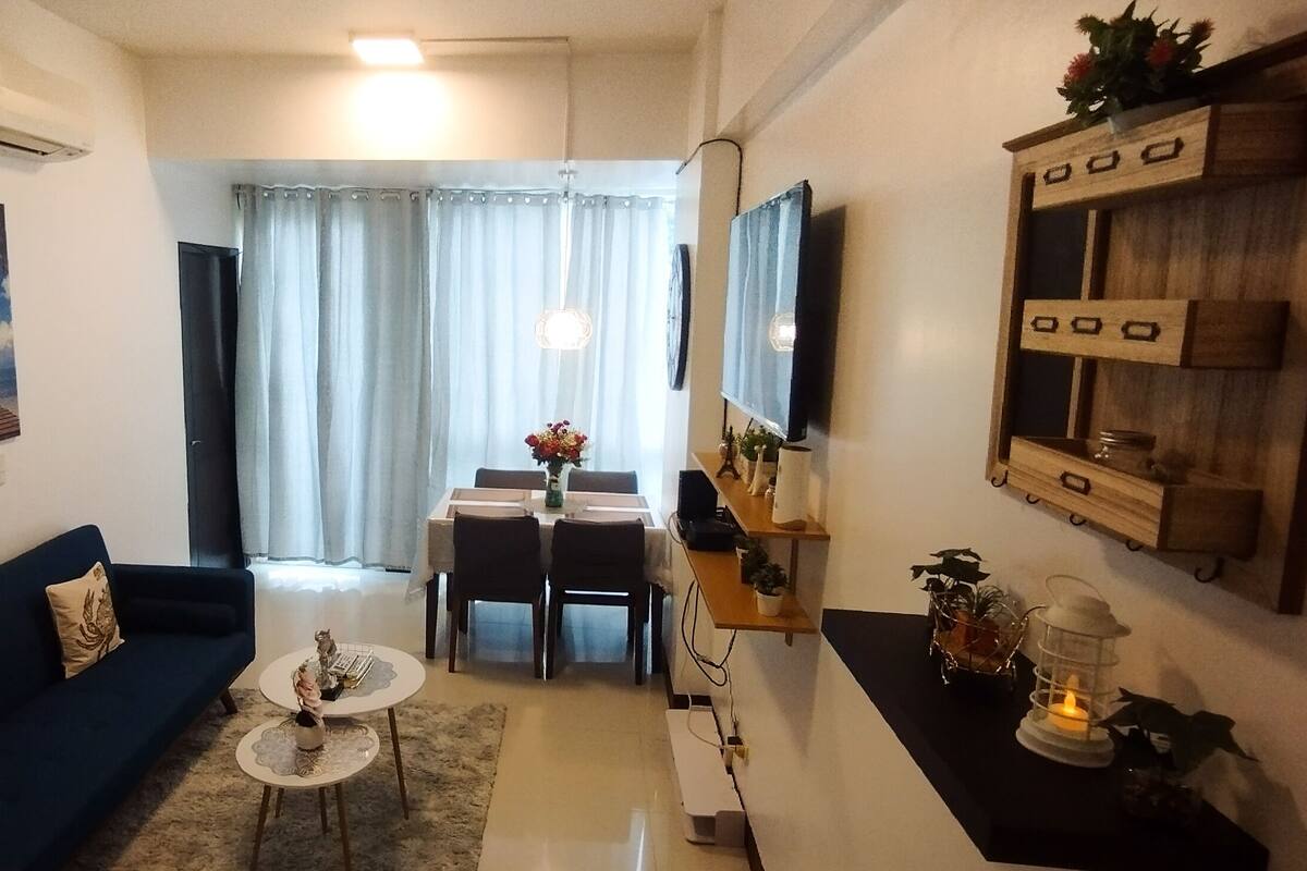 A compact living space with a dark blue sofa, a dining table with chairs, a TV mounted on the wall, and a small side table with a lantern.