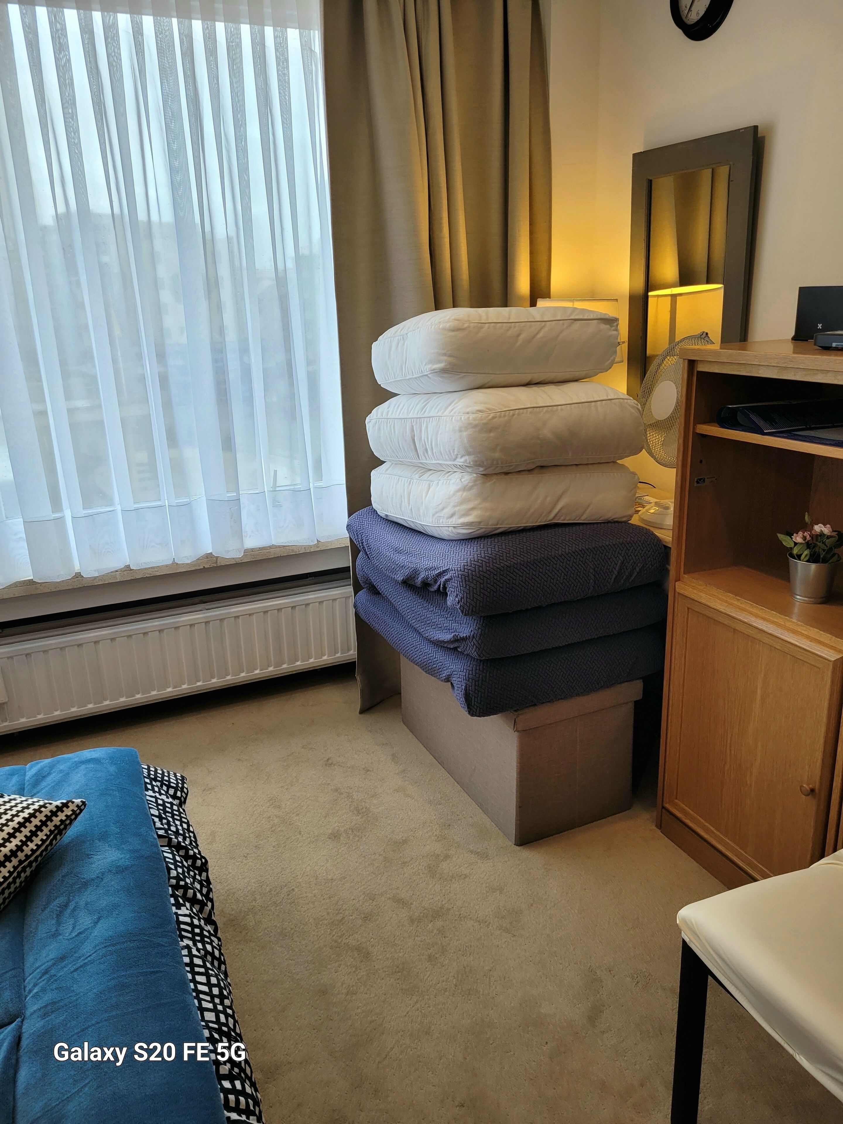 Une chambre d’hôtel avec un lit, une table de chevet avec une lampe, un radiateur, une fenêtre avec des rideaux et une pile d’oreillers.