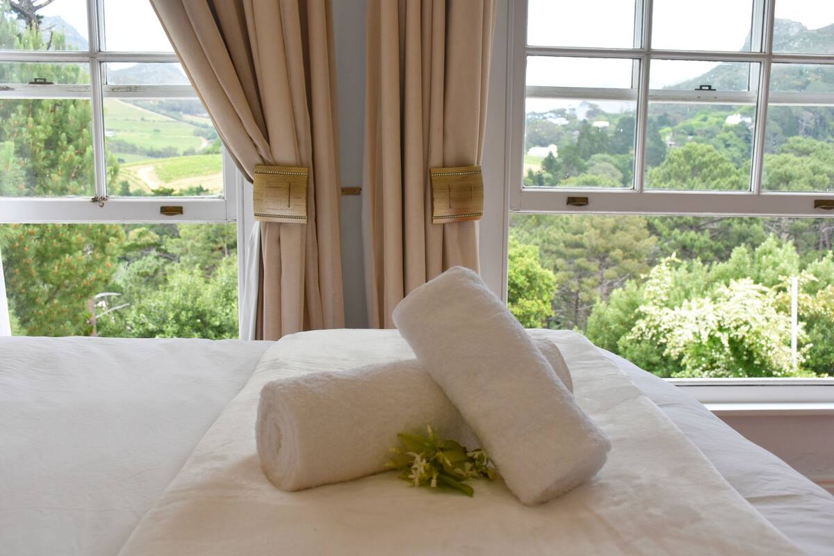 A bed with white linens and a rolled towel, a window with beige curtains, and a view of greenery outside.
