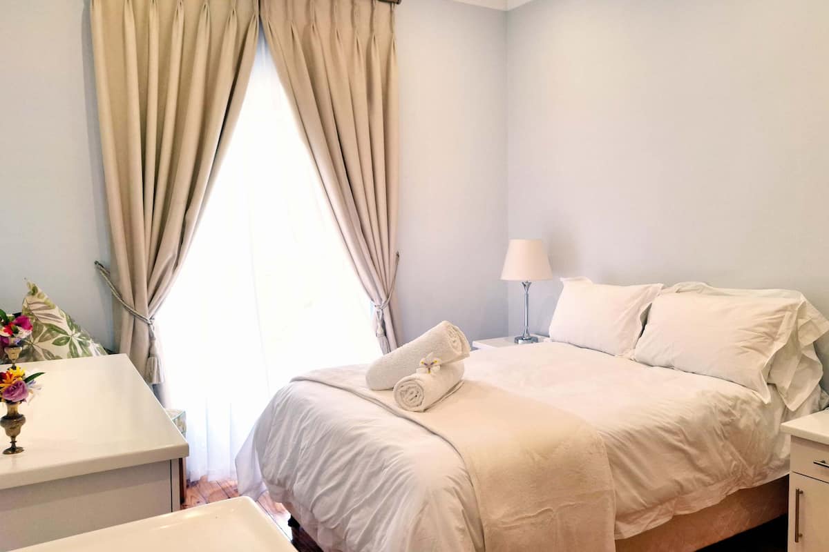 A hotel room with a bed, two bedside tables, a nightstand, a lamp, a vase with flowers, and a window with curtains.