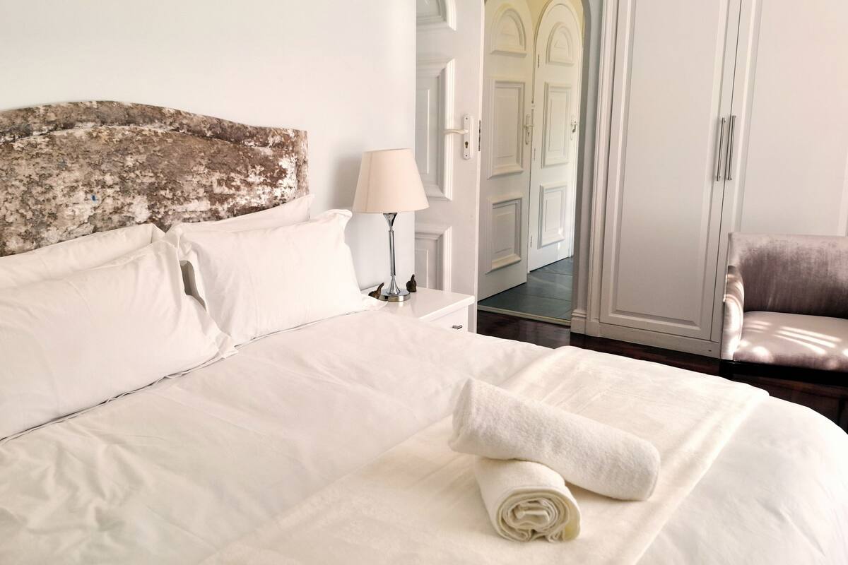 A neatly made bed with white linens, a lamp on a bedside table, and a chair in the background.