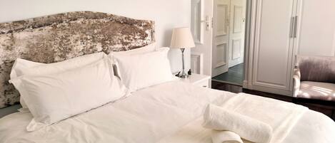 A neatly made bed with white linens, a lamp on a bedside table, and a chair in the background.