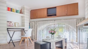 Un salon moderne avec un bureau en bois, une chaise blanche et un pouf noir. Un téléviseur est fixé au-dessus du bureau, sur des meubles en bois.