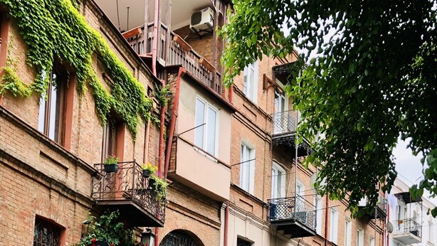 Une rue pavée bordée d’immeubles à plusieurs étages, avec des balcons ornés de plantes et des garde-corps en fer forgé.