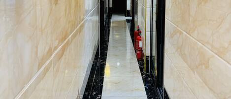 Un pasillo con paredes de mármol y suelo de baldosas blanco y negro, equipado con lámparas de pared y un extintor.