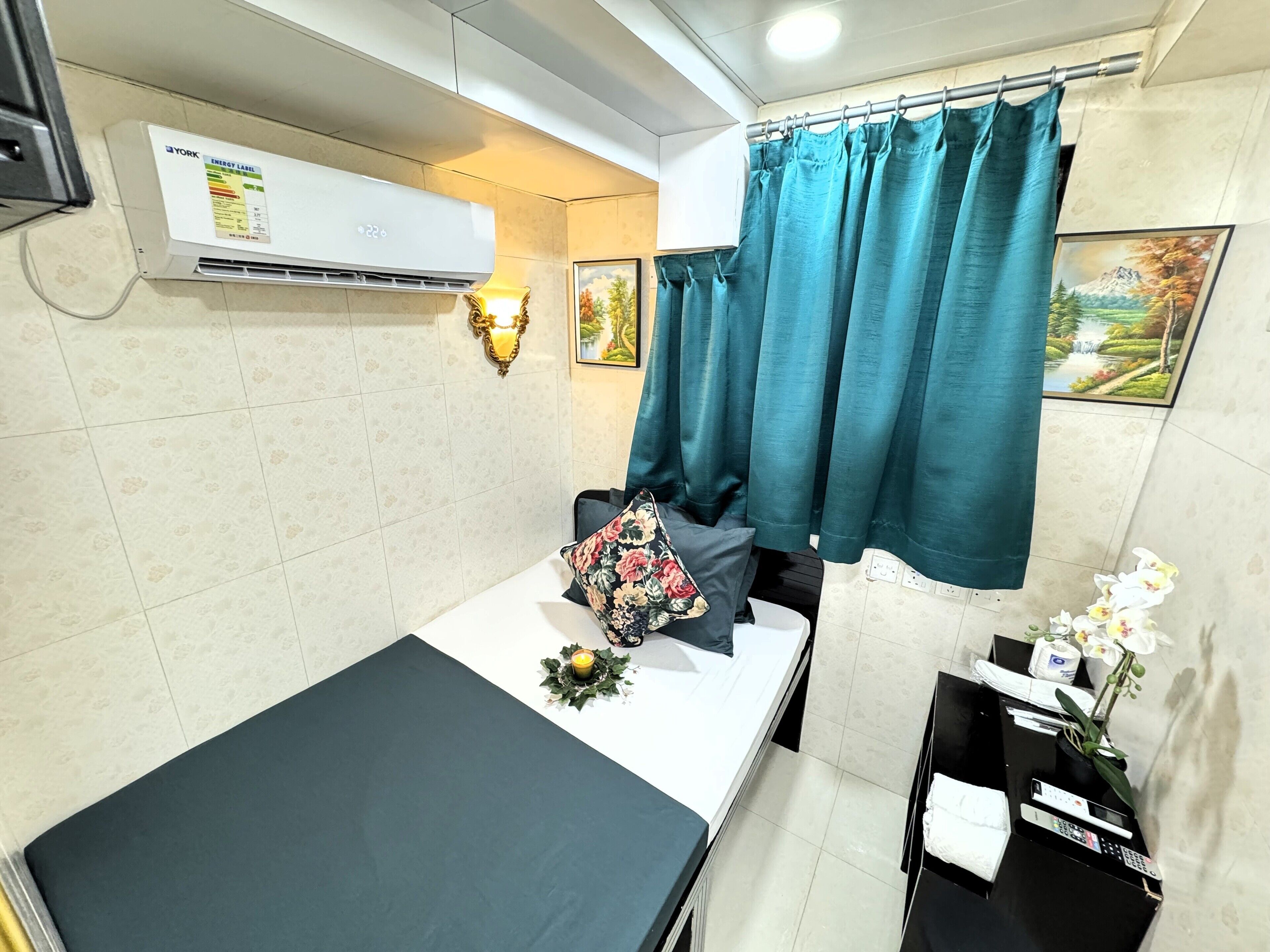 A hotel room with a bed, a small table with a remote control, a wall-mounted air conditioner, teal curtains, and a wall-mounted light fixture.