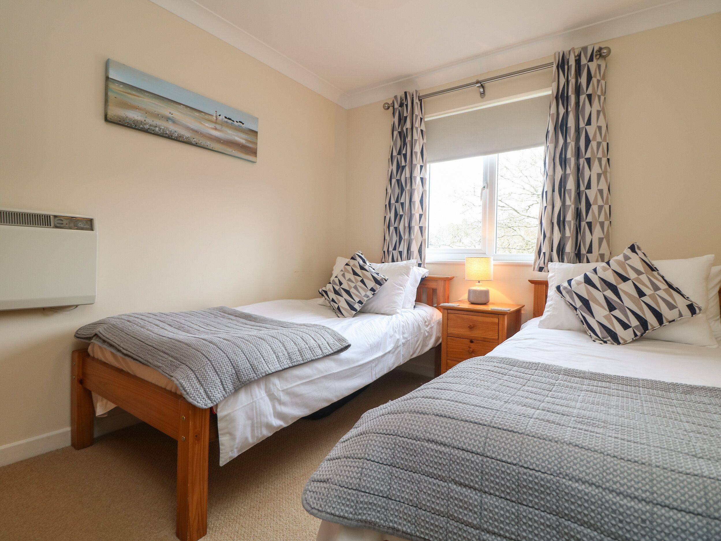 A bedroom with two single beds, a bedside table with a lamp, a painting on the wall, and a window with patterned curtains.