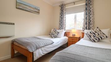 A bedroom with two single beds, a bedside table with a lamp, a painting on the wall, and a window with patterned curtains.